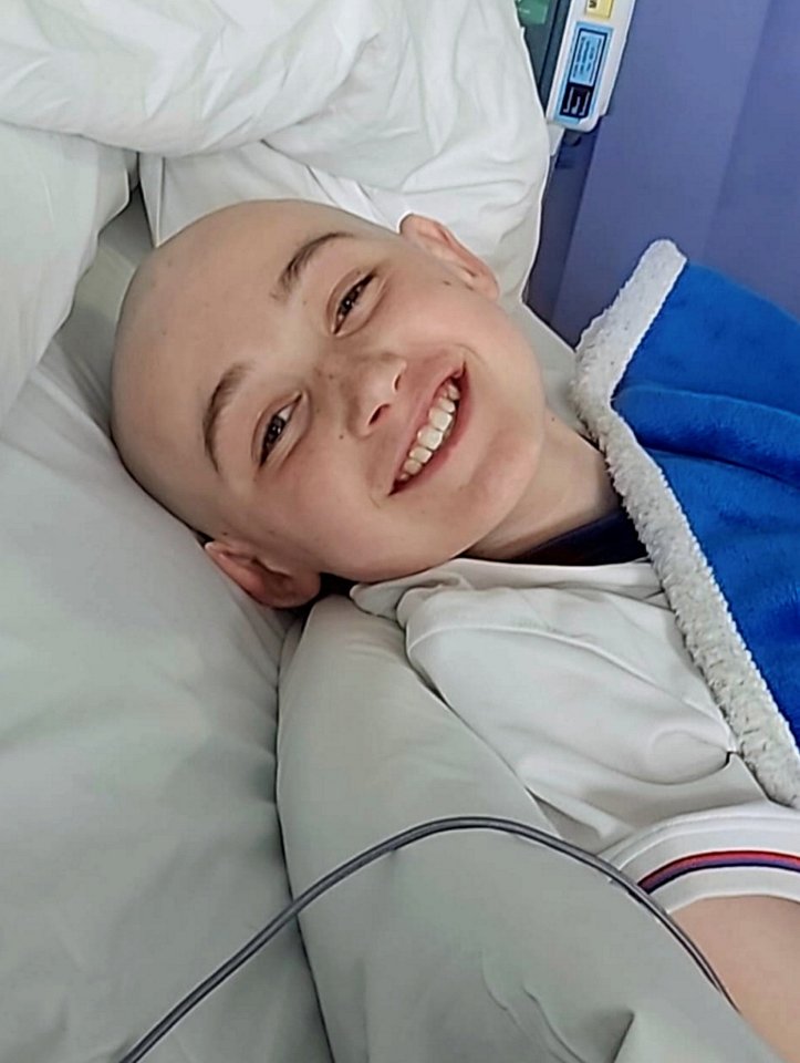 Smiling boy with shaved head lying in hospital bed.