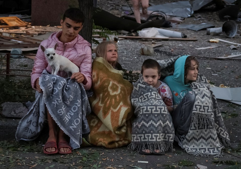 Residents wrapped in blankets sit amidst the rubble of a bombed-out apartment building.