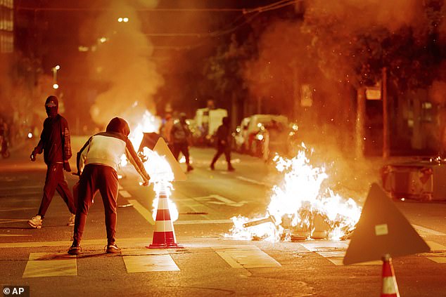 A person sets a traffic cone on fire as burning containers block the street during the third night of riots in Lausanne, Switzerland, Tuesday August 26, 2025