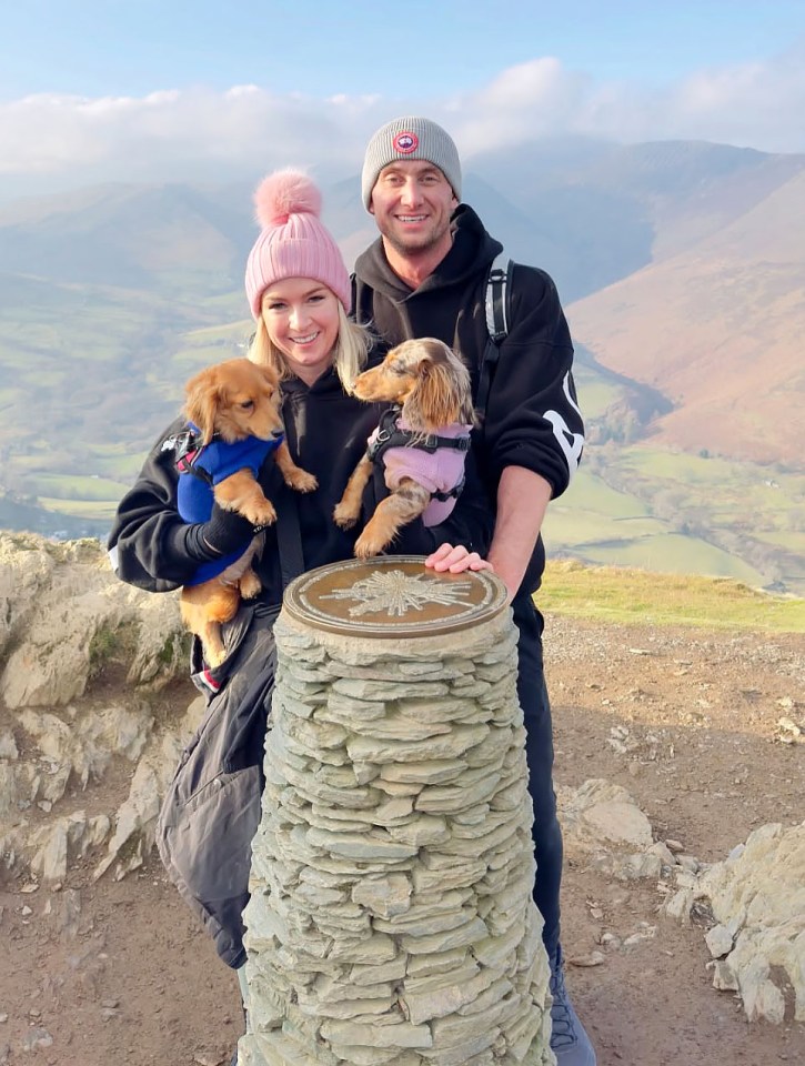 Couple with two small dogs posing by a stone marker on a mountaintop.