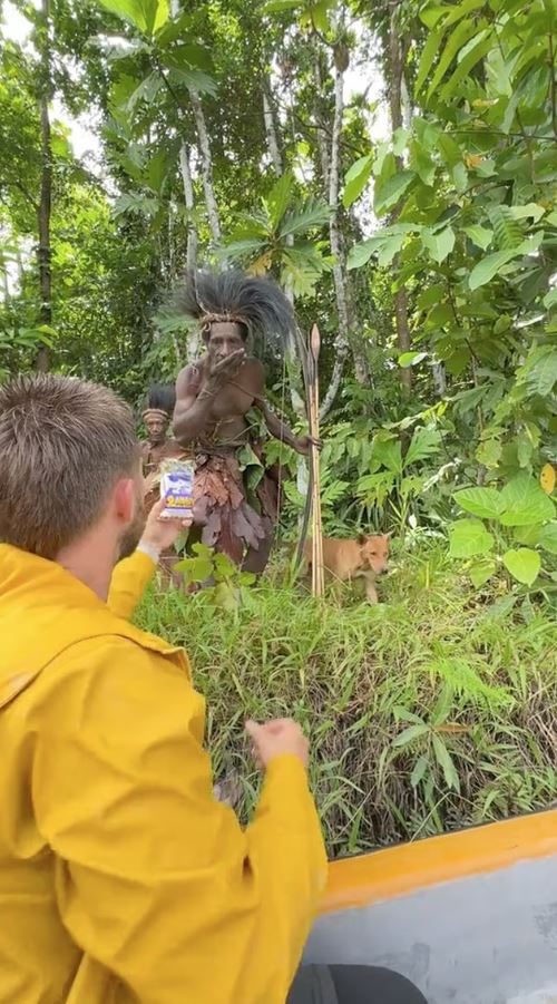 A man in a yellow raincoat offers a package to a man in traditional tribal attire holding a spear.