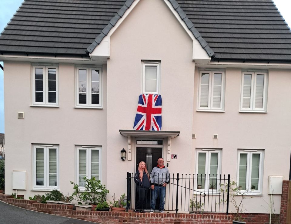 A couple stands outside their new home, which has a British flag displayed.