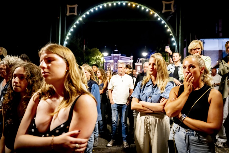 A crowd of people standing at night, appearing concerned.