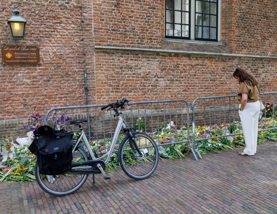 A woman stands by a bike and floral tributes outside a church.
