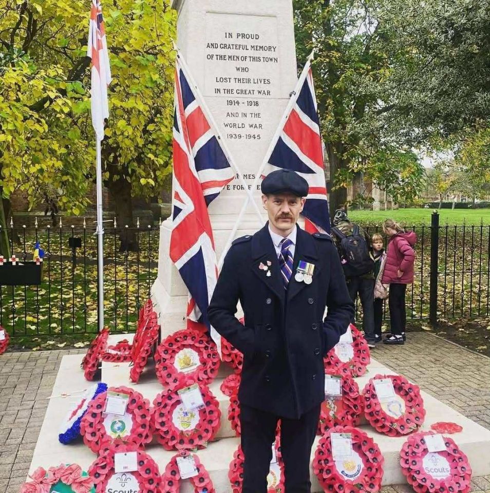Man in military-style coat standing by a war memorial with wreaths.