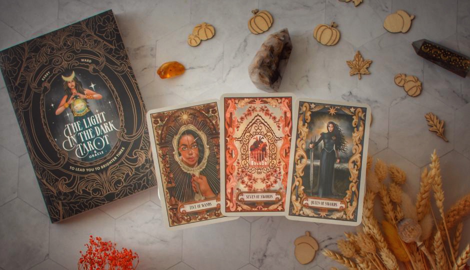 Three tarot cards (Five of Wands, Seven of Swords, Queen of Swords) and "The Light in the Dark Tarot & Oracle" book.