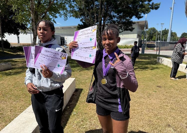 Two girls holding certificates and a medal outdoors.