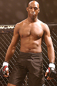 A muscular man with a shaved head stands in a fighting cage.
