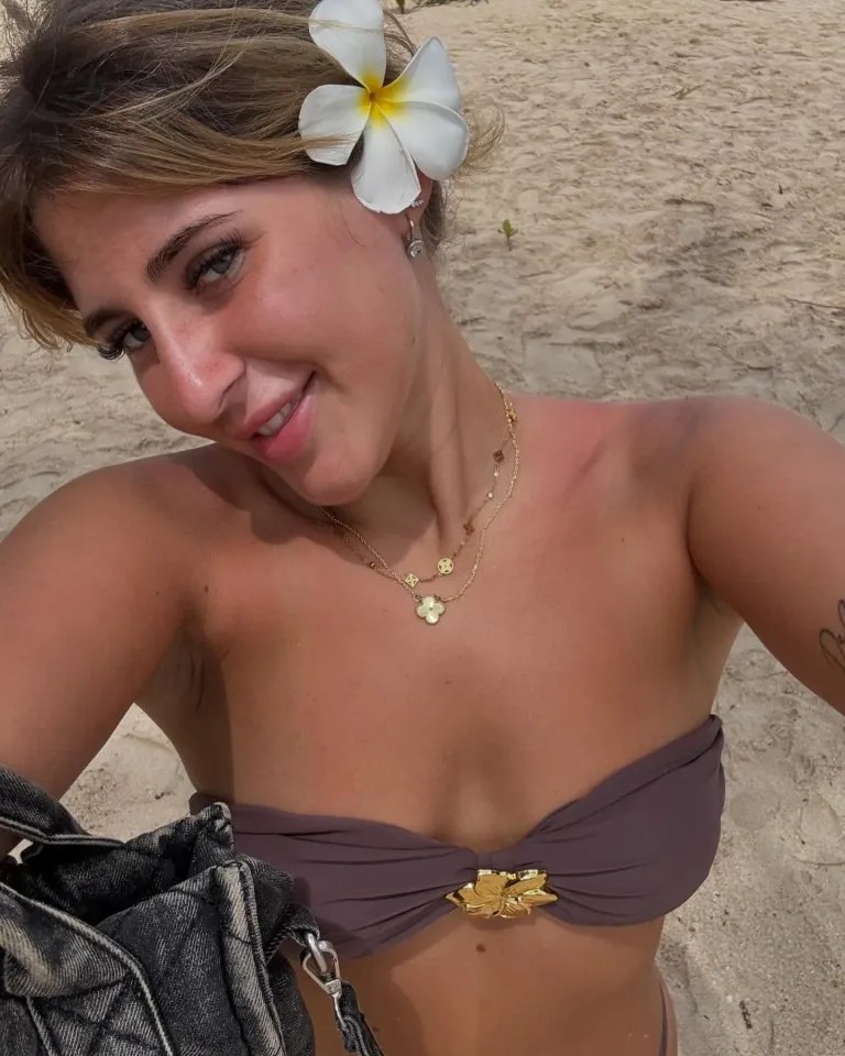 Woman on a beach wearing a purple bikini top and gold necklaces.