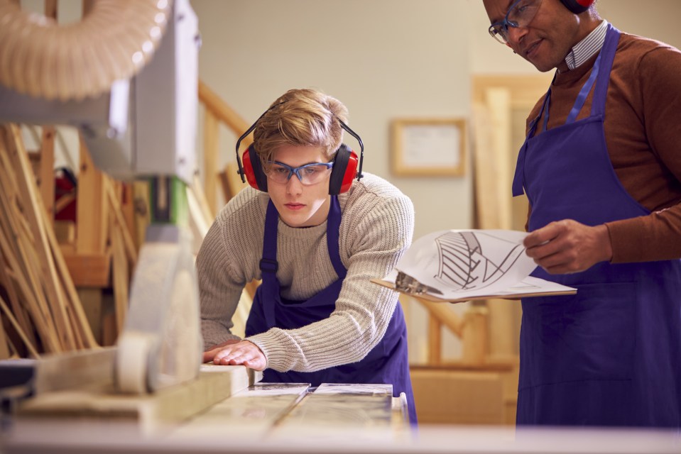 A carpentry student uses a bench saw while his tutor looks on.