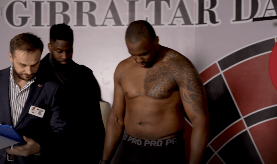 A shirtless man with tattoos stands being weighed, while two men in suits watch.