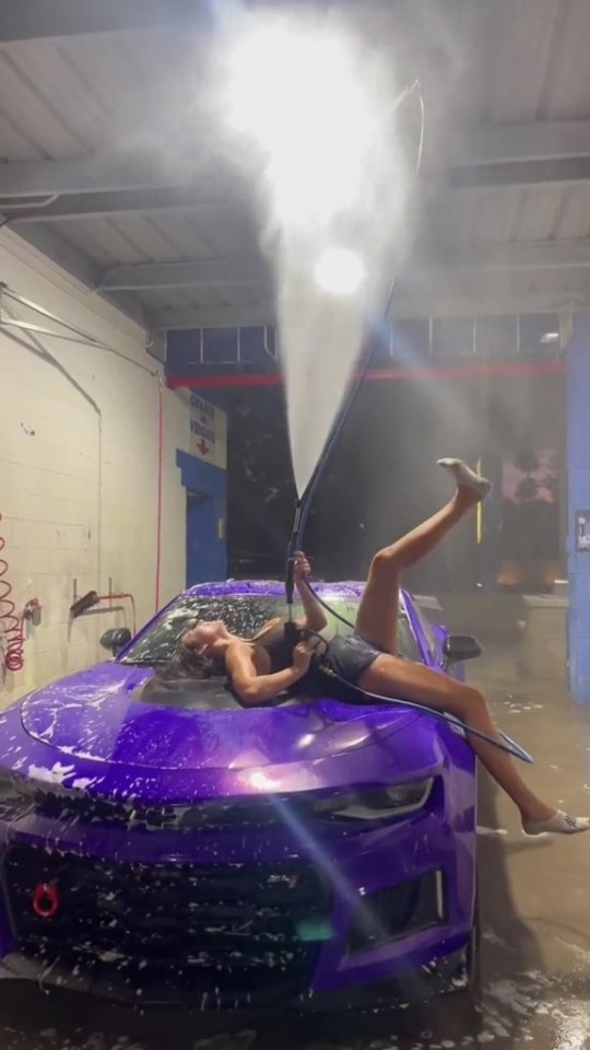Woman lying on a purple car being sprayed with a high-pressure hose.