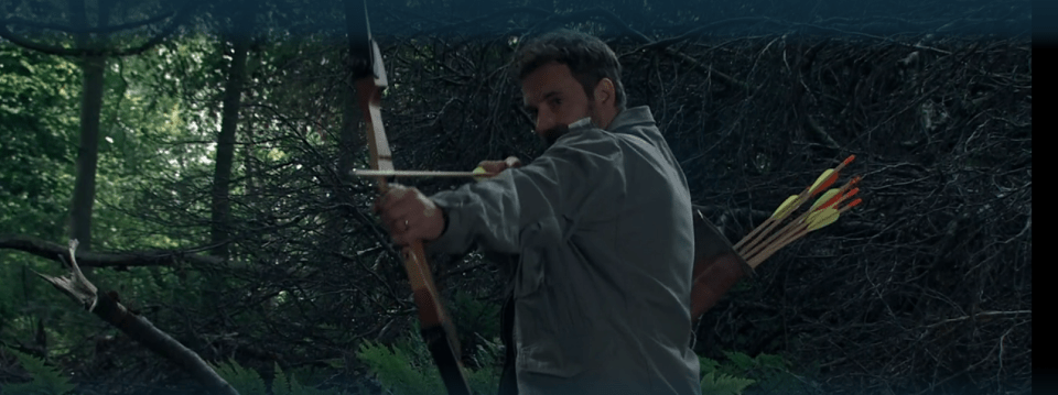 Man aiming a bow and arrow in a forest.