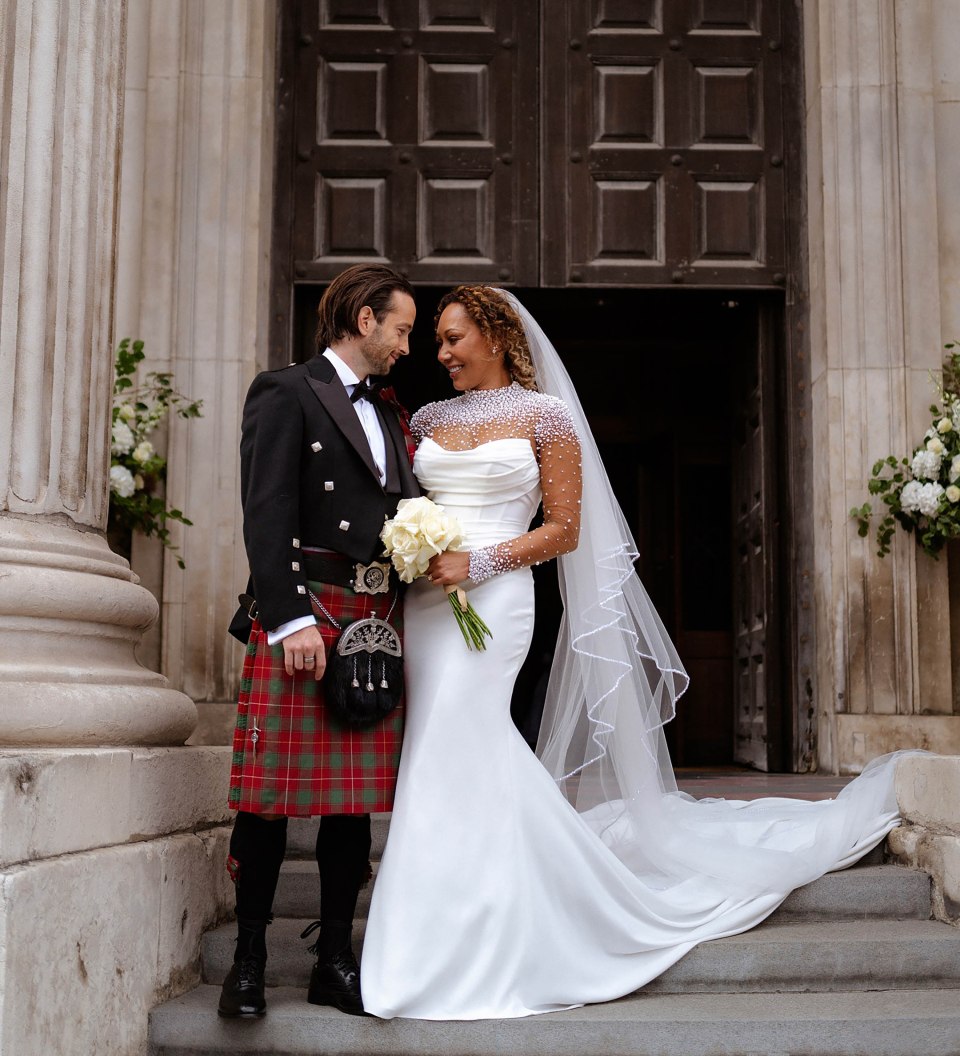Newlyweds in wedding attire stand on steps outside a building.