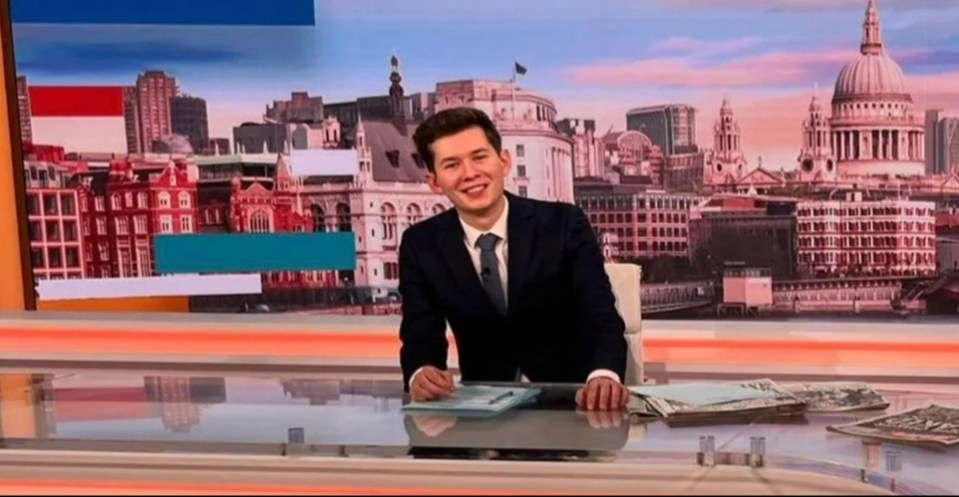 A news presenter sits at a desk in a television studio with a London cityscape backdrop.