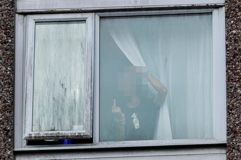An asylum seeker gives the middle finger to protesters through a hotel window.