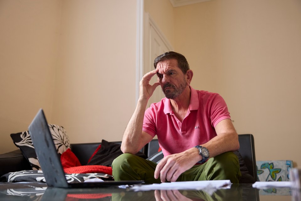 A man sits on a couch looking stressed while looking at a laptop and paperwork.
