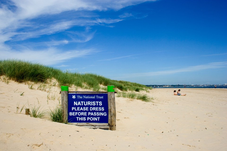 Naturists: Please dress before passing this point sign on a sandy beach.