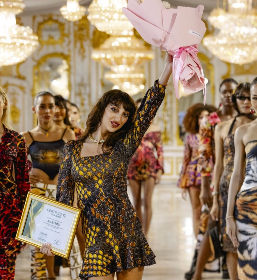 Woman in snakeskin-patterned dress holding award and flowers after fashion show.