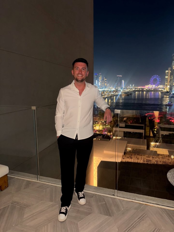 Man in white shirt and black pants standing on balcony overlooking city at night.