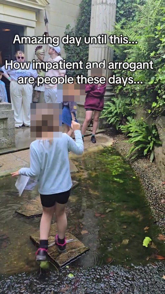 A child crossing stepping stones in a shallow pool of water, with impatient adults waiting nearby.