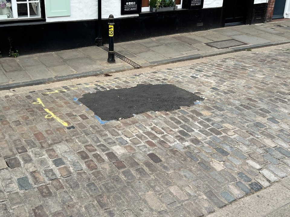 Tarmac repair patch on a cobblestone street in front of buildings.