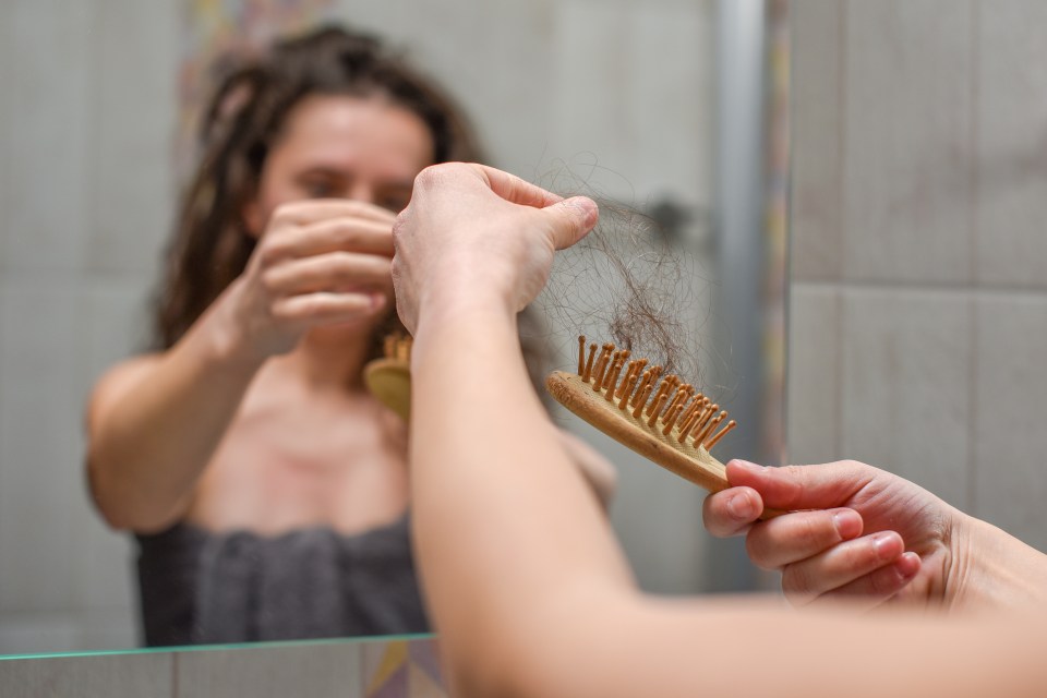 Woman looking at a brush full of hair in a bathroom mirror.