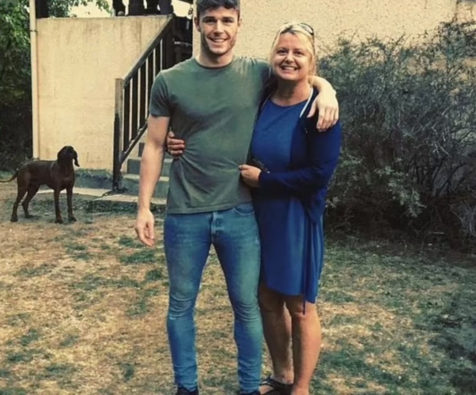 A man and woman stand together outdoors, arm in arm; a dog is visible in the background.