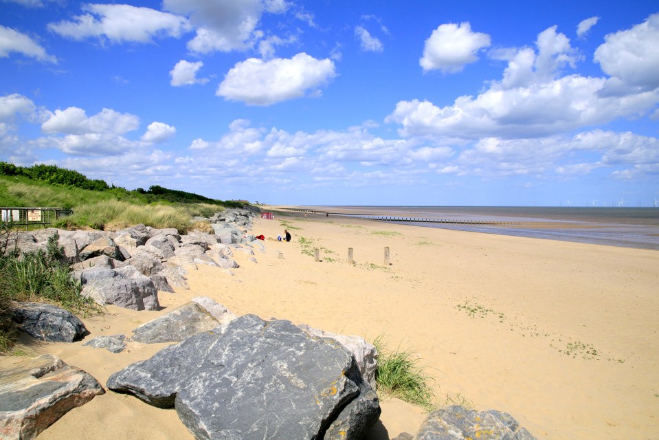 Sandy beach with rocks, people, and wind turbines in the distance.