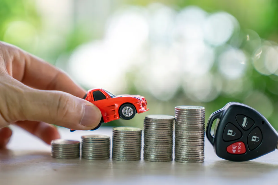 A red toy car about to be placed on stacks of coins next to a car key.