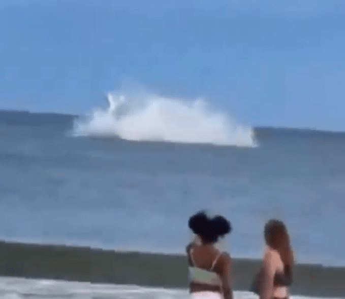 Two people on a beach watch a large wave break in the ocean.