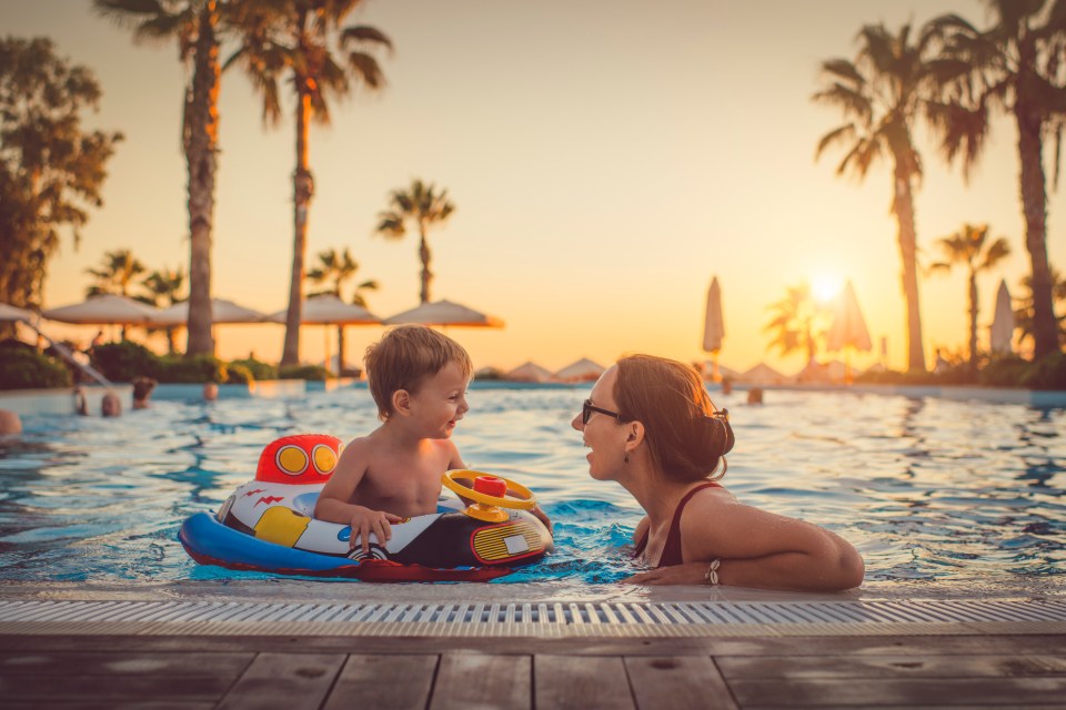 A mother and her toddler son enjoy a summer vacation, playing in a resort pool at sunset.