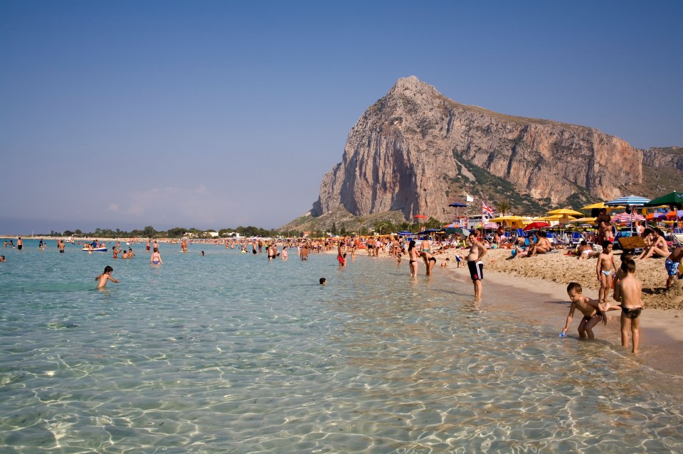 Crowded beach with clear water and a large rock in the background.