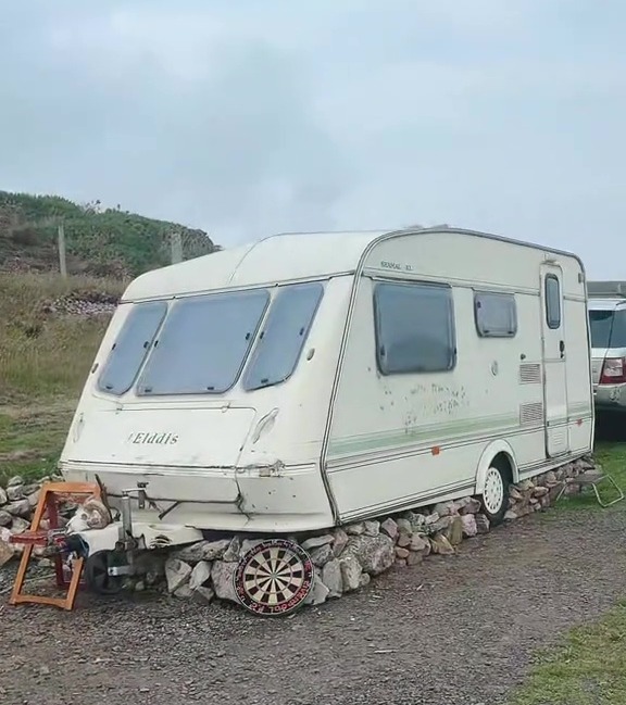 Caravan parked on a stone base with a dartboard in front.