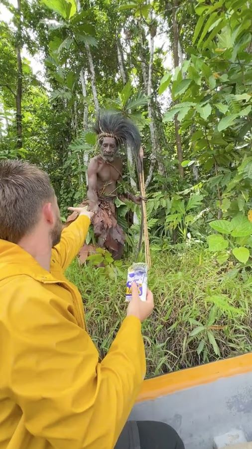 A man in a yellow jacket offers a package to a tribesman in a jungle.