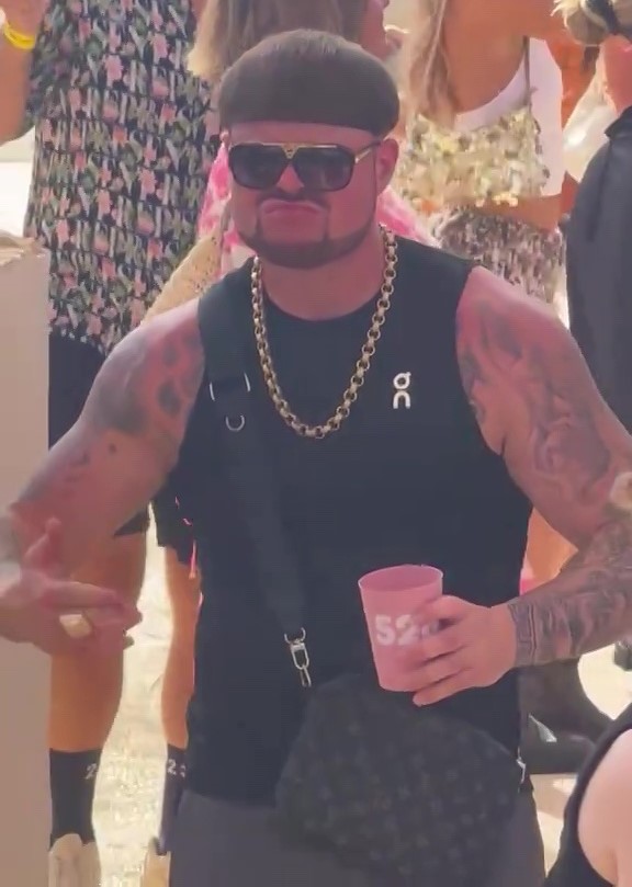 A man with a bowl cut, sunglasses, gold chain, and tattoos holds a pink cup at a party.