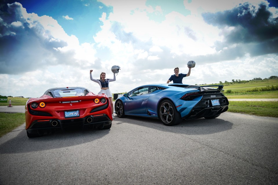 Two people holding helmets stand triumphantly between a red Ferrari and a blue Lamborghini.