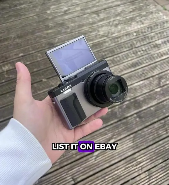 A hand holding a silver Lumix camera with its screen flipped up.