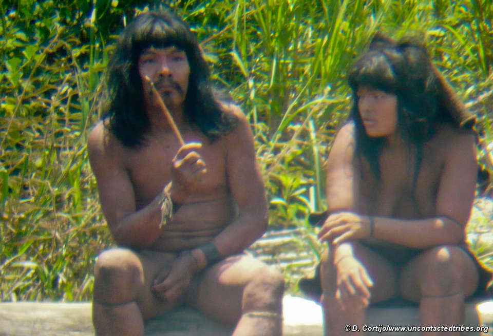 Two Mashco-Piro people sitting in the jungle.  One holds a knife.