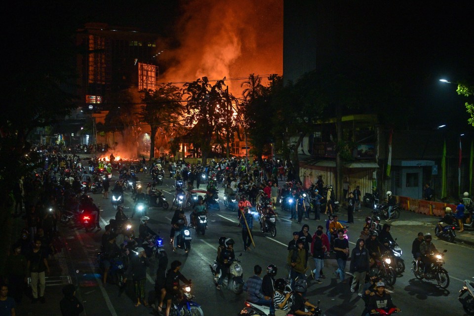 Protesters on motorcycles at night near a burning building.