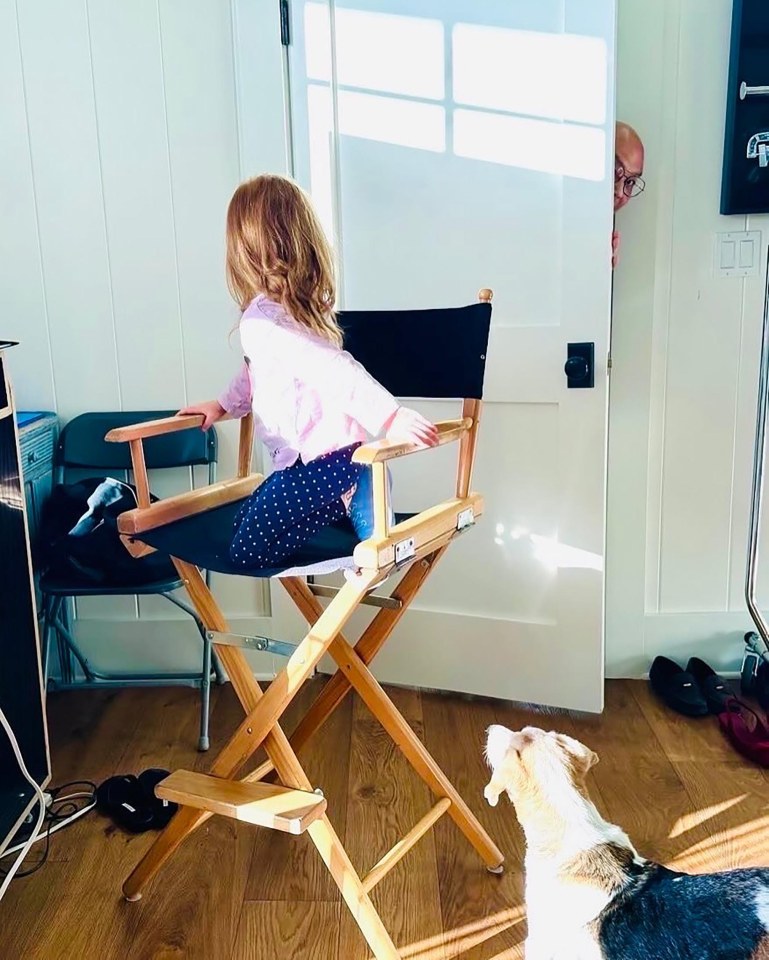 A toddler sits in a director's chair; a man peeks from behind a door; a dog looks on.