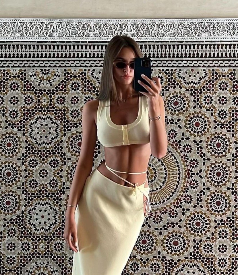 Woman in a pale yellow crop top and skirt taking a selfie in front of an ornate wall.