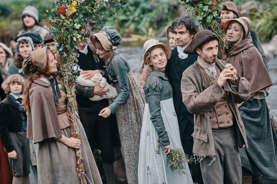 A crowd of people in period clothing gather outdoors; some carry flowering branches.
