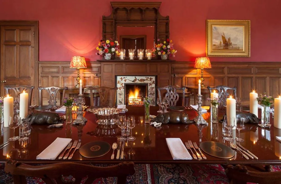 A formally set dining table in a wood-paneled room with a fireplace.