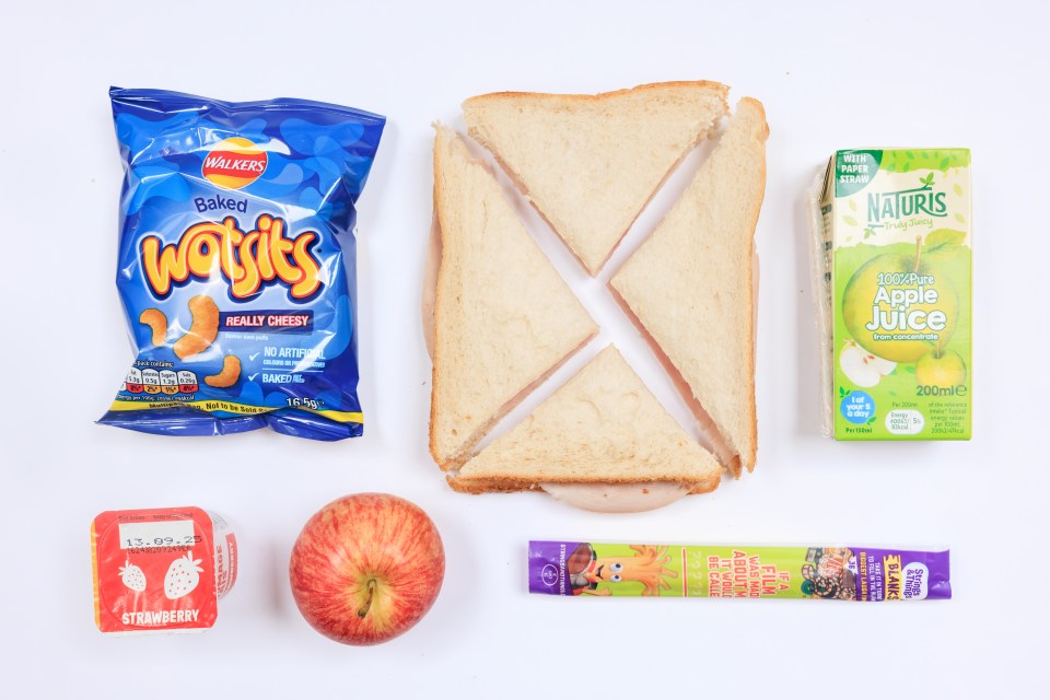 A packed lunch containing a ham sandwich, apple, apple juice, yogurt, cheese stick, and a bag of crisps.