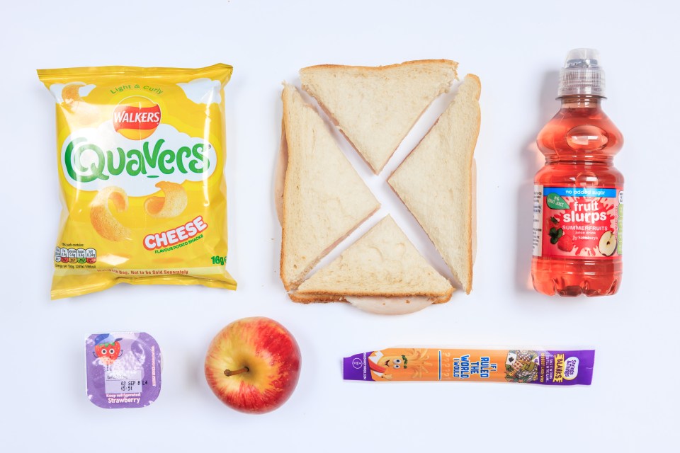 Photo of a packed lunch containing a sandwich, crisps, fruit juice, apple, yogurt and cheese stick.