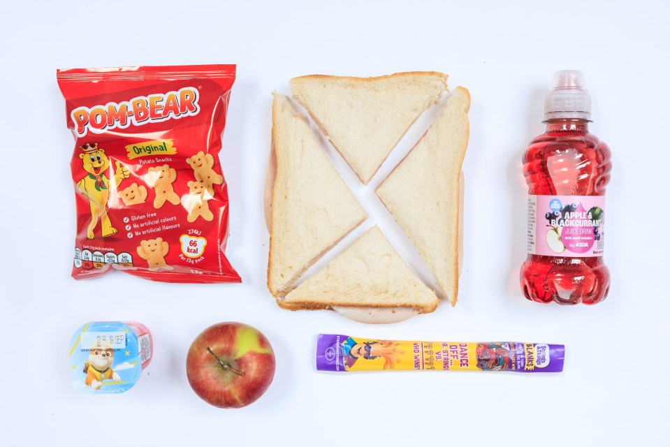 Photo of a packed lunch containing a ham sandwich, Pom-Bear potato snacks, an apple, yogurt, and apple & blackcurrant juice.