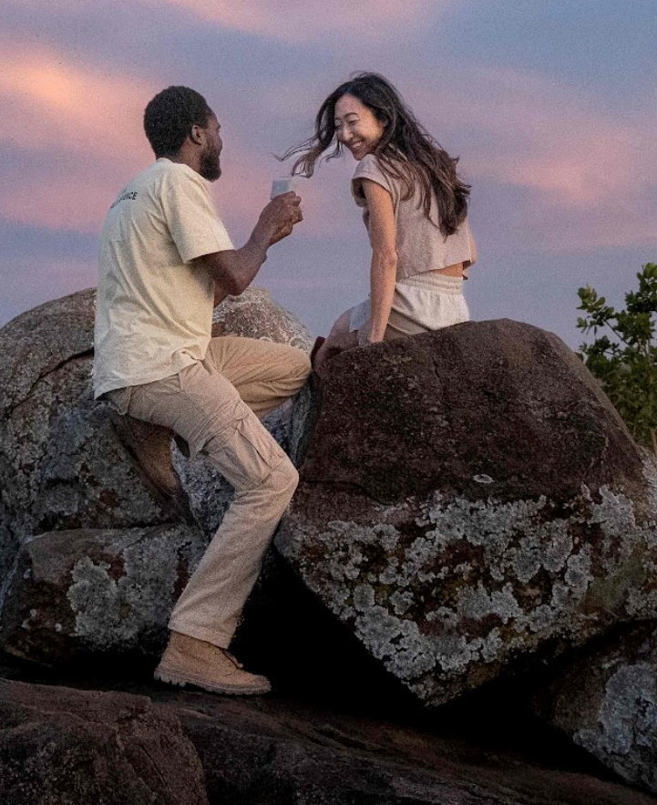 A man and woman sharing a drink while sitting on rocks at sunset.