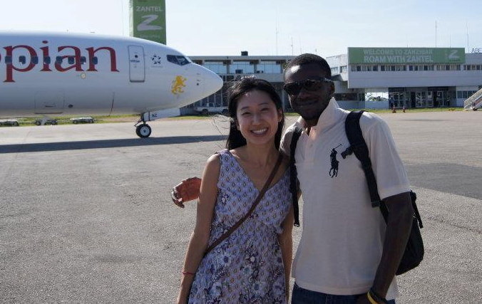 A woman and a man stand together at an airport, smiling for a photo in front of an airplane.