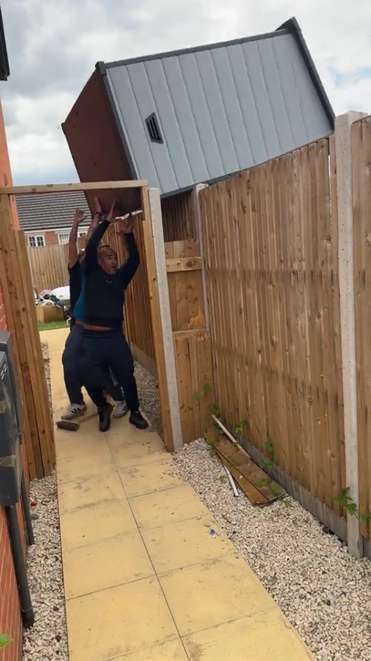 Two people struggling to move a garden shed through a narrow gate.